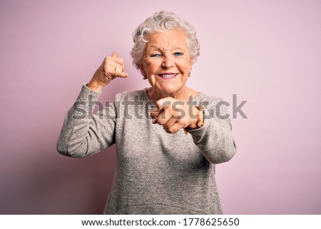 Senior beautiful woman wearing casual t-shirt standing over isolated pink background smiling doing talking on the telephone gesture and pointing to you. Call me.