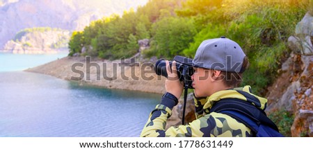 Teenager boy holding digital camera and shooting landscape with lake in the mountains in summer day.