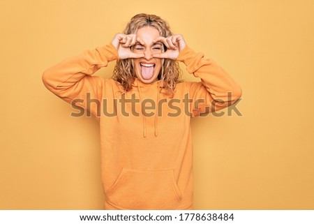 Young beautiful blonde sporty woman wearing casual sweatshirt over yellow background doing ok gesture like binoculars sticking tongue out, eyes looking through fingers. Crazy expression.