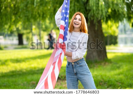 Young red haired woman holding USA national flag standing outdoors in summer park. Positive girl celebrating United States independence day.