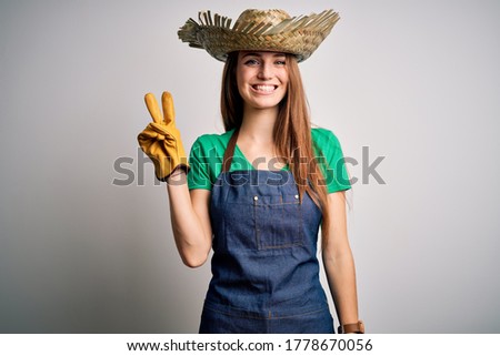 Young beautiful redhead farmer woman wearing apron and hat over white background smiling with happy face winking at the camera doing victory sign. Number two.