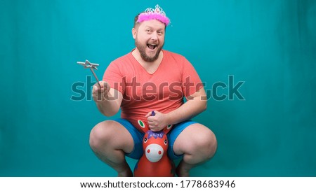handsome bearded freaky man in a pink T-shirt with a diadem on his head is riding astride a unicorn with a magic wand in his hand. A funny wizard joke to make and fulfill a wish