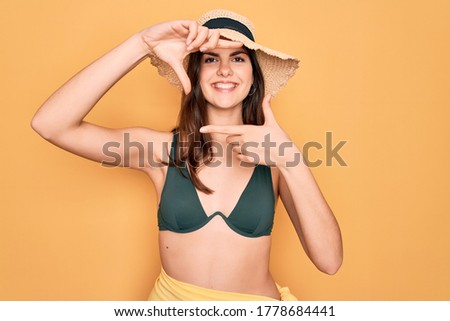Young beautiful girl wearing swimwear bikini and summer sun hat over yellow background smiling making frame with hands and fingers with happy face. Creativity and photography concept.