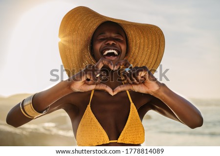 Smiling woman in bikini standing on the beach making a heart shape with her fingers. Female at the seashore wearing a sun hat making heart with hands.
