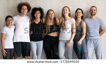 Group portrait of smiling beautiful multiracial diverse young people in sportswear embracing posing near wall, feeling excited of yoga morning routine workout in modern club, looking at camera. Royalty-Free Stock Photo #1778849030