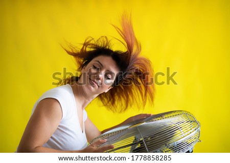 A beautiful red-haired woman is cooled off standing over a large electric fan on a yellow background. Girl with hair developing in the wind. Device for cooling the air.