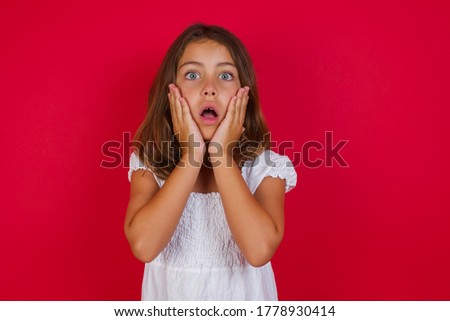 Studio shot of scared terrified Little caucasian girl witth blue eyes wearing white dress standing over isolated red background . customer shocked with prices at shop, being short of money 