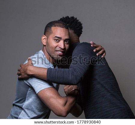 Two adult athletic black men hugging, in the sport clothes, isolated on a warm grey background, one man is looking in the camera