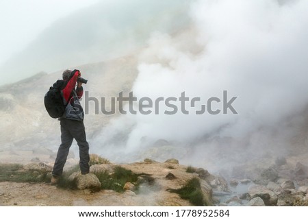 Male photographer takes pictures of mysterious volcanic landscape, aggressive hot spring, eruption fumarole, gas-steam activity in crater active volcano. Mount landscape, travel destinations for hike.