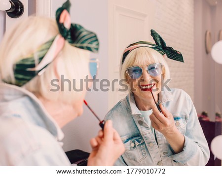 Smiling elderly senior stylish woman in blue sunglasses and denim jacket using red lipstick by the mirror in stylish loft interior. Style, fashion, make up, anti age concept Royalty-Free Stock Photo #1779010772
