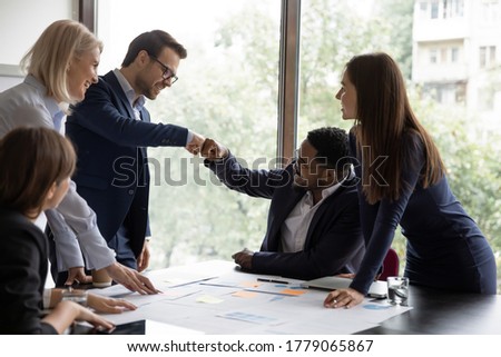 Multi ethnic businessmen project leaders fists bump celebrating corporate achievements, workmates meet in office boardroom surrounded by happy workmates. Reach common decision, share advance concept Royalty-Free Stock Photo #1779065867