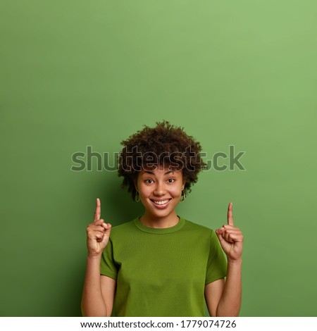 Positive dark skinned woman shows copy space above, points index fingers upwards, demonstrates nice advertisement, suggests good idea, isolated on green background. People and promotion concept