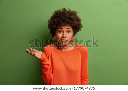 Doubtful indecisive woman raises palm with hesitation, faces difficult question or two choices, wears orange sweater and earrings, isolated on green background. People, perception and attitude Royalty-Free Stock Photo #1779074975