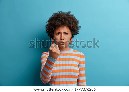 Disturbed angry woman clenches fist and has outraged expression, warns about revenge, expresses hate and negative emotions, wants punch enemy, dressed casually, isolated on blue studio wall. Royalty-Free Stock Photo #1779076286