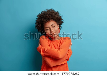 Self care and tenderness concept. Pleased dark skinned woman embraces herself, holds shoulders, feels comfort in her new sweater, has romantic tender mood, needs feel warmth, comfort and love Royalty-Free Stock Photo #1779076289