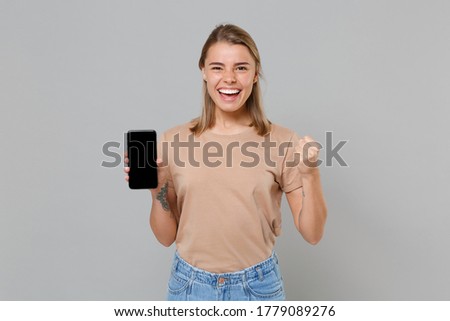 Joyful young blonde woman girl in casual beige t-shirt isolated on gray wall background. People lifestyle concept. Mock up copy space. Hold mobile phone with blank empty screen, doing winner gesture