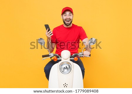 Delivery man in red cap t-shirt uniform driving moped motorbike scooter hold mobile phone isolated on yellow background studio Guy employee working courier Service quarantine pandemic covid19 concept