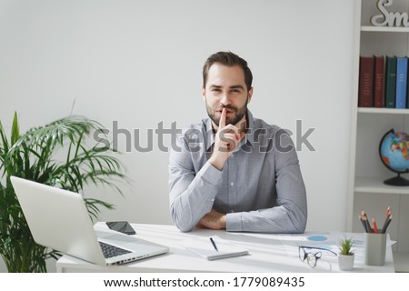 Secret young business man in gray shirt sit at desk work on laptop in light office on white wall background. Achievement business career concept. Saying hush be quiet with finger on lips shhh gesture