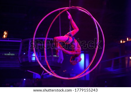 Flexible young woman make performance on aerial hoop, flexible back on aerial hoop, aerial circus show, purple red light. Flexible woman gymnast upside down on hoop. Night club performance