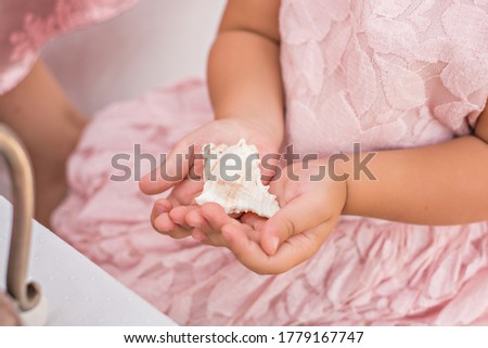 A little cute blonde girl in a gentle pink dress holds a seashell in her hands by the sea. A beautiful baby girl plays with sea treasures. Decoration of celebrations, photo zones in elegant marine 