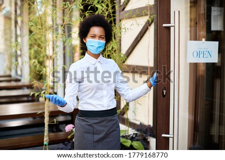 Happy African American cafe owner wearing protective face mask while standing at entrance door and reopening after coronavirus epidemic. 