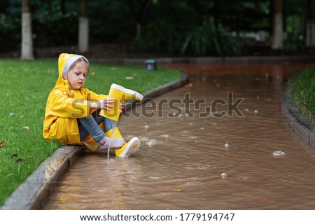 Cute little girl in yellow raincoat and rubber boots walking outdoor during rain. Bad weather, summer tropical storm, autumn fashion concept. Royalty-Free Stock Photo #1779194747