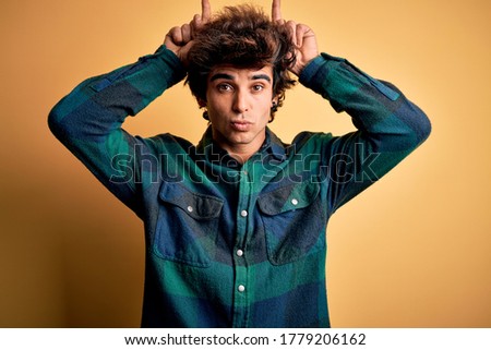Young handsome man wearing casual shirt standing over isolated yellow background doing funny gesture with finger over head as bull horns