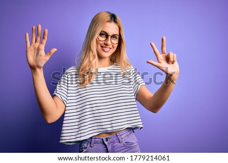 Young beautiful blonde woman wearing striped t-shirt and glasses over purple background showing and pointing up with fingers number eight while smiling confident and happy.