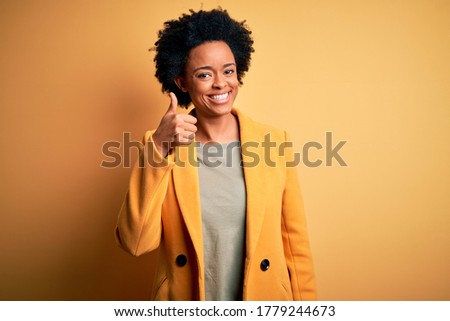 Young beautiful African American afro businesswoman with curly hair wearing yellow jacket doing happy thumbs up gesture with hand. Approving expression looking at the camera showing success.