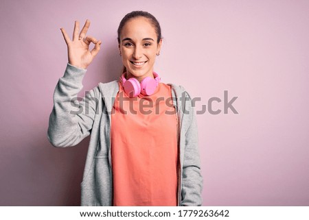 Beautiful sporty woman doing sport listening to music using headphones over pink background smiling positive doing ok sign with hand and fingers. Successful expression.