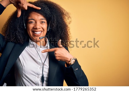 Young african american operator woman with afro hair wearing headset over yellow background smiling making frame with hands and fingers with happy face. Creativity and photography concept.