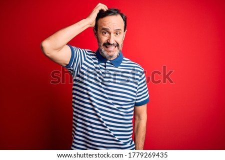 Middle age hoary man wearing casual striped polo standing over isolated red background confuse and wonder about question. Uncertain with doubt, thinking with hand on head. Pensive concept.