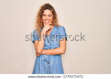 Young beautiful woman with blue eyes wearing casual denim dress over white background looking confident at the camera smiling with crossed arms and hand raised on chin. Thinking positive.
