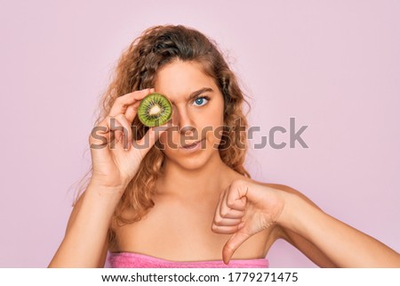 Beautiful woman with blue eyes wearing towel shower after bath holding kiwi fruit over eye with angry face, negative sign showing dislike with thumbs down, rejection concept