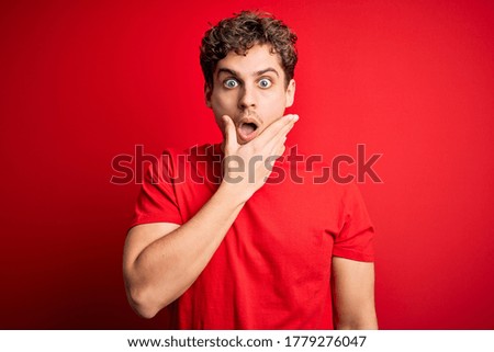 Young blond handsome man with curly hair wearing casual t-shirt over red background Looking fascinated with disbelief, surprise and amazed expression with hands on chin