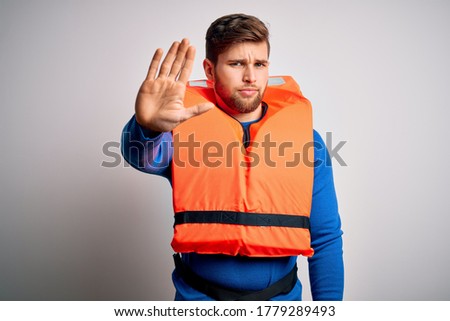 Young blond tourist man with beard and blue eyes wearing lifejacket over white background doing stop sing with palm of the hand. Warning expression with negative and serious gesture on the face.