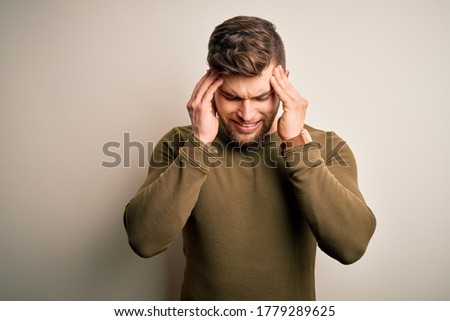 Young blond man with beard and blue eyes wearing green sweater over white background with hand on head for pain in head because stress. Suffering migraine.