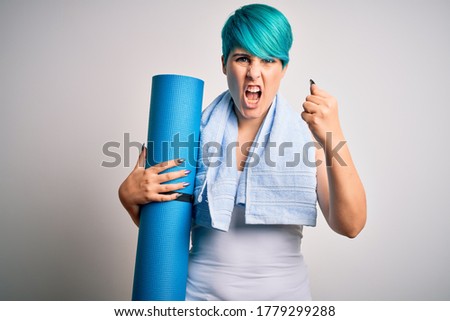Young sporty woman with blue fashion hair holding mat after yoga over yellow background annoyed and frustrated shouting with anger, crazy and yelling with raised hand, anger concept