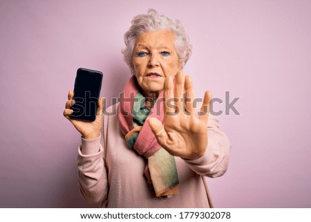 Senior beautiful grey-haired woman holding smartphone showing screen over pink background with open hand doing stop sign with serious and confident expression, defense gesture
