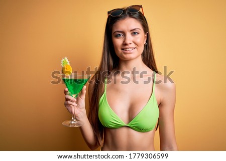 Young beautiful woman with blue eyes on vacation wearing bikini drinking cocktail with a happy face standing and smiling with a confident smile showing teeth