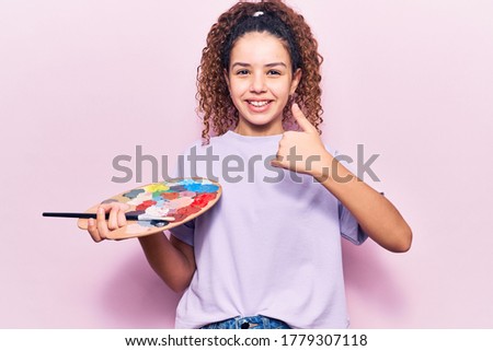 Beautiful kid girl with curly hair holding paintbrush and palette smiling happy and positive, thumb up doing excellent and approval sign 