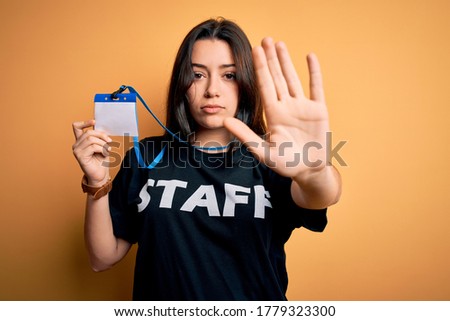 Young brunette worker woman wearing staff t-shirt as uniform showing id card with open hand doing stop sign with serious and confident expression, defense gesture