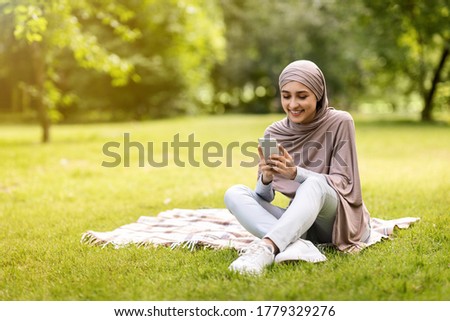 Cheerful muslim woman looking at smartphone screen, sitting on grass at park, having lovely weekend, copy space