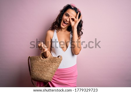 Young beautiful woman with curly hair on vacation wearing swimsuit holding beach bag with happy face smiling doing ok sign with hand on eye looking through fingers