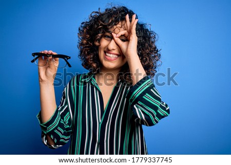 Young beautiful optical arab woman holding vision glasses over isolated blue background with happy face smiling doing ok sign with hand on eye looking through fingers