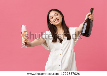 Celebration, party holidays and fun concept. Happy smiling asian woman host of event, holding bottle champagne and glass and reaching hands for hugging guest, standing pink background