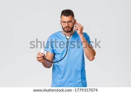 Healthcare workers, medicine, covid-19 and pandemic self-quarantine concept. Serious-looking focused young doctor, physician in ER wearing scrubs, glasses, listening patient lungs with stethoscope