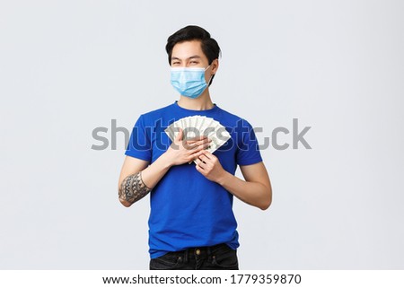 Money, lifestyle, insurance and investment concept. Satisfied griedy asian guy hugging cash of dollars and smiling pleased, wearing medical mask, earned or won lottery