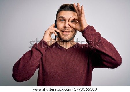 Young man with blue eyes speaking on the phone having a conversation on smartphone with happy face smiling doing ok sign with hand on eye looking through fingers