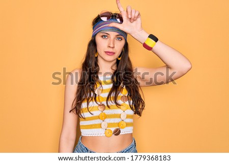 Young beautiful hippie woman with blue eyes wearing accesories and sunnglasses making fun of people with fingers on forehead doing loser gesture mocking and insulting.